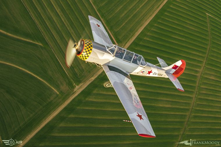 Yak aerobatic trainer ZU-YAK flying over green farmland, air-to-air photograph by Frank Moody